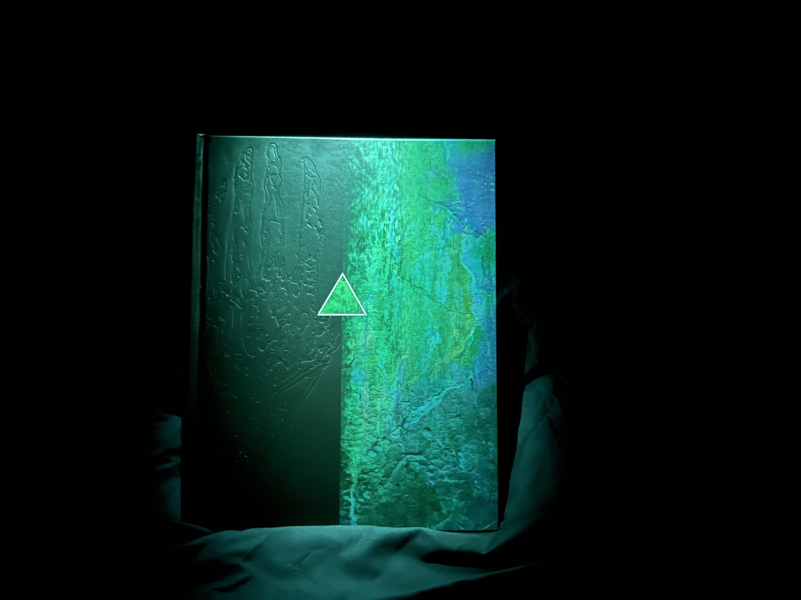 a black and green copy of Delta Green: The Conspiracy sits in the darkness illuminated only by a green spotlight from above. Strange, alien symbols are pressed into the cover.