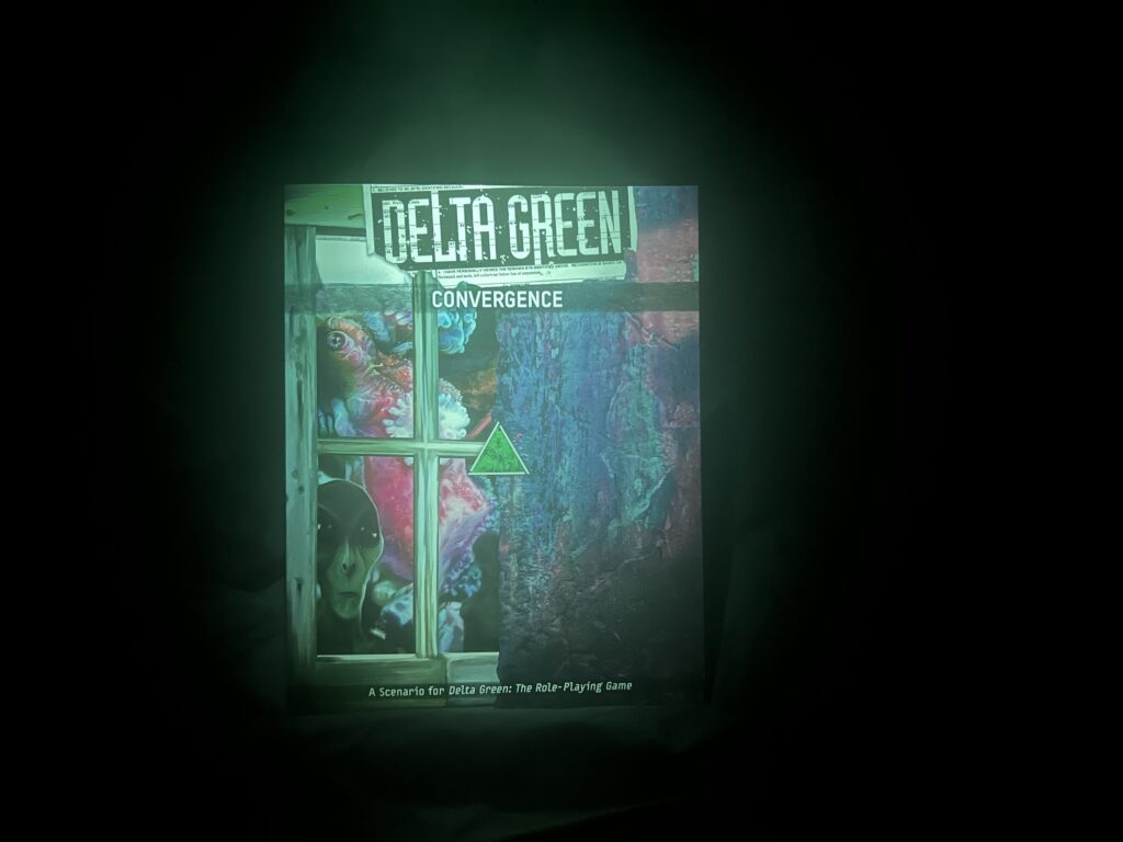 A paperback copy of Delta Green: Convergence sits in the smokey darkness illuminated only by a green light from above. The cover of the book shows a pale grey alien peering into a window